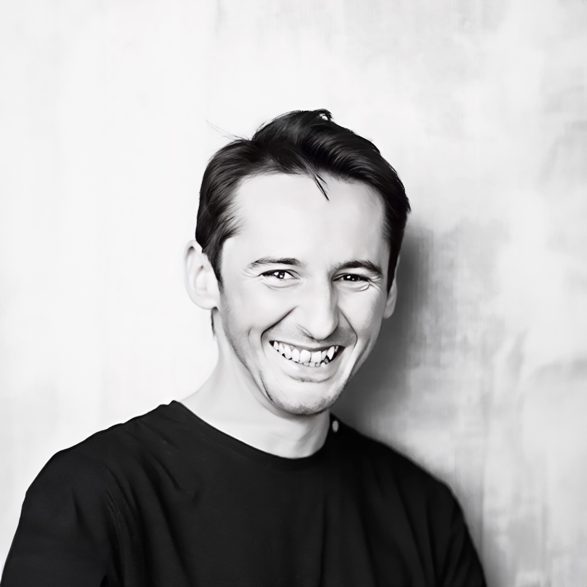 Porträt von Denis, einem Meisterfriseur und Kollegen im Salon, mit einem warmen, einladenden Lächeln und einem stilvollen Haarschnitt, in einem schwarzen Hemd vor einem strukturierten grauen Hintergrund.
