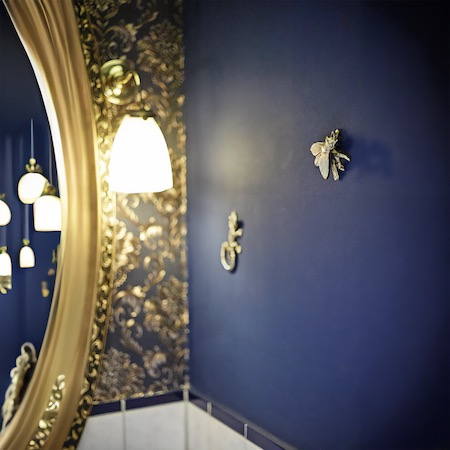 Interieur des Salons mit einer eleganten goldverzierten Spiegelrahmung, klassischer Wandbeleuchtung und dekorativen Elementen an einer dunkelblauen Wand.